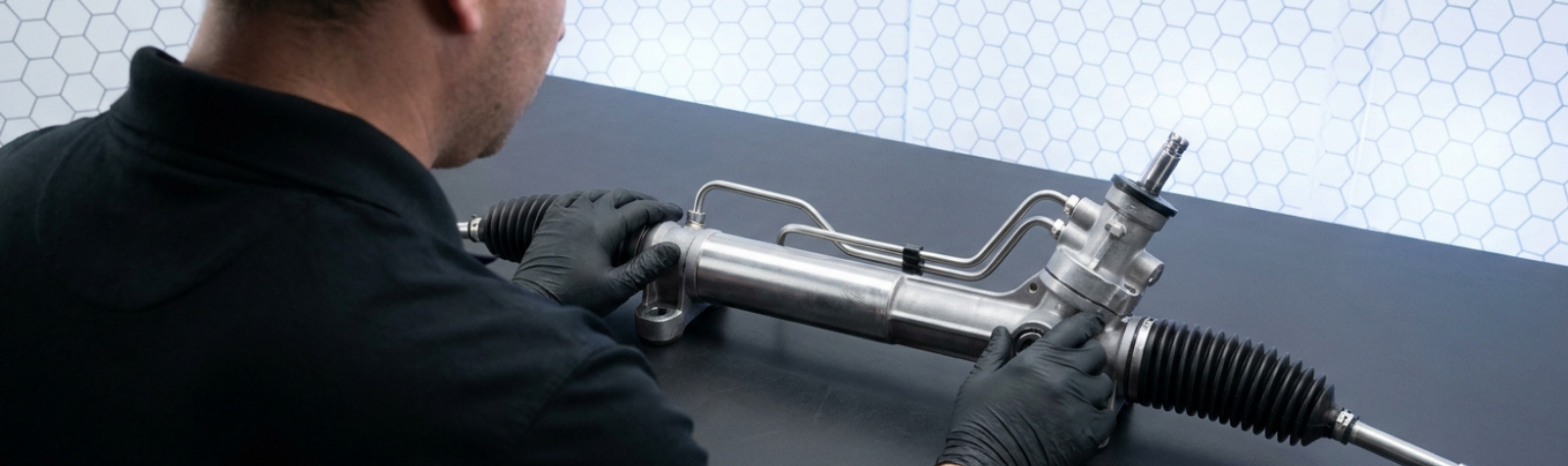 Técnico examinando una cremallera de dirección hidráulica con latiguillos y fuelles sobre una mesa de trabajo.